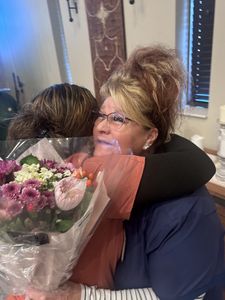 One adult holding a bouquet of flowers embraces another adult indoors