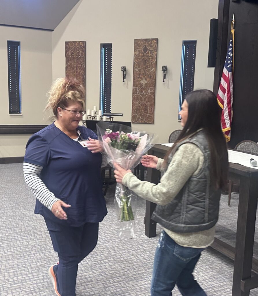 One adult hands a bouquet of flowers to another indoors during a Caregiver of the Year award presentation.