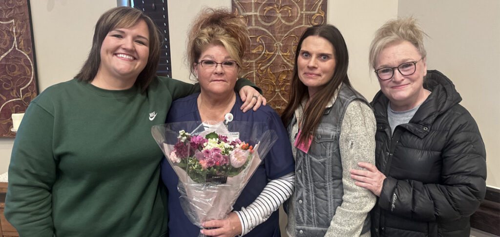 Group of four adults indoors, with the center person holding flowers while being recognized as Caregiver of the Year.
