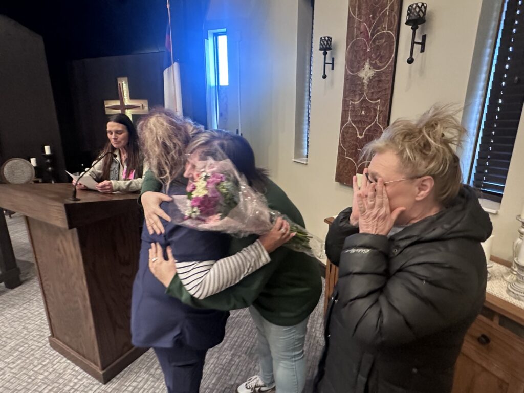Caregiver of the Year recipient receives hugs while holding flowers during an indoor recognition event.